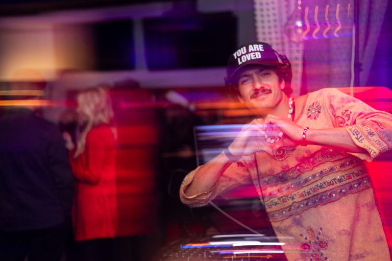 Scott Welch in a YOU ARE LOVED cap making a heart with his hands behind the decks